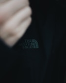 A close-up of a black jacket featuring 'The North Face' logo prominently displayed in white stitching. The image includes part of a blurred hand in the frame, creating a sense of movement or adjustment.