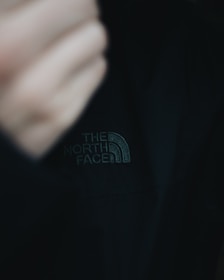 A close-up of a black jacket featuring 'The North Face' logo prominently displayed in white stitching. The image includes part of a blurred hand in the frame, creating a sense of movement or adjustment.