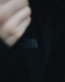 A close-up of a black jacket featuring 'The North Face' logo prominently displayed in white stitching. The image includes part of a blurred hand in the frame, creating a sense of movement or adjustment.