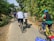 A group of people are cycling along a narrow, paved rural road surrounded by lush green foliage and intermittent sunlight. The riders are wearing casual attire and helmets, with one of them on a bright yellow bicycle. The path is lined with bamboo and other greenery, suggesting a peaceful and scenic environment.