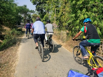 A group of people are cycling along a narrow, paved rural road surrounded by lush green foliage and intermittent sunlight. The riders are wearing casual attire and helmets, with one of them on a bright yellow bicycle. The path is lined with bamboo and other greenery, suggesting a peaceful and scenic environment.