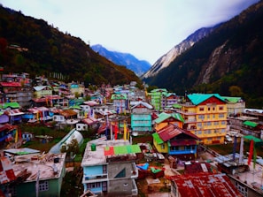 A vibrant village nestled in a mountainous valley features an array of colorful houses with bright roofs and walls. The surrounding landscape includes lush green forests and towering steep mountains under a cloudy sky. Prayer flags are visible throughout, adding to the cultural ambiance.