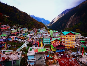 A vibrant village nestled in a mountainous valley features an array of colorful houses with bright roofs and walls. The surrounding landscape includes lush green forests and towering steep mountains under a cloudy sky. Prayer flags are visible throughout, adding to the cultural ambiance.