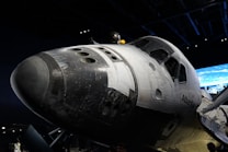 A space shuttle is prominently displayed, showcasing its black and white thermal tiles and windows. The backdrop includes a dimly lit room with a screen displaying an Earth view.