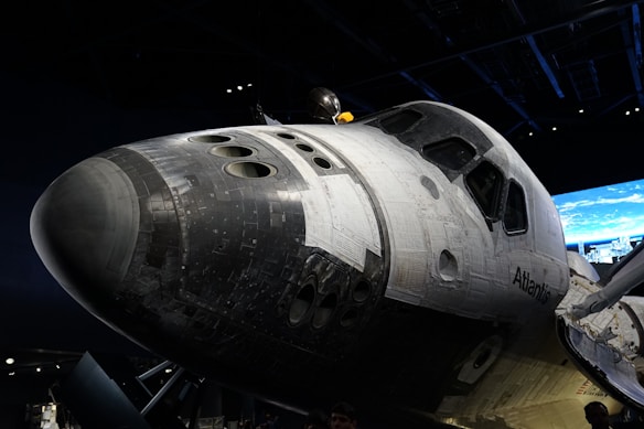 A space shuttle is prominently displayed, showcasing its black and white thermal tiles and windows. The backdrop includes a dimly lit room with a screen displaying an Earth view.