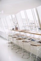 A modern, minimalist interior space featuring a long wooden bar with several high stools underneath. The bar is adorned with a chess set, some books, and a small plant, placed near large, angled windows that allow natural light to flood the area. There is a sleek, geometrically designed floor lamp near the left side of the bar.