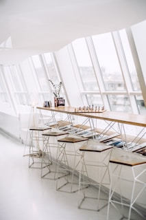 A modern, minimalist interior space featuring a long wooden bar with several high stools underneath. The bar is adorned with a chess set, some books, and a small plant, placed near large, angled windows that allow natural light to flood the area. There is a sleek, geometrically designed floor lamp near the left side of the bar.