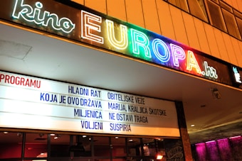A cinema entrance with a colorful neon sign displaying the name 'Kino Europa'. Below the sign is a marquee listing movie titles in a foreign language.