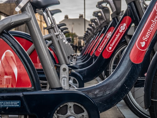 A close-up perspective of a row of bicycles available for hire in a city bike-sharing scheme. The bikes have a sturdy build with a prominent red branding label visible on the rear wheel covers. The gray bike frames and large wheels are lined up symmetrically, suggesting a well-organized docking system.