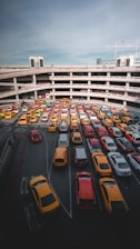 A multi-level parking garage filled with rows of brightly colored cars, predominantly yellow and red. The structure's concrete layers create a pattern with the cars neatly aligned along the lanes. The sky above is clear with a few wispy clouds and some construction cranes visible in the background.