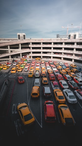 A multi-level parking garage filled with rows of brightly colored cars, predominantly yellow and red. The structure's concrete layers create a pattern with the cars neatly aligned along the lanes. The sky above is clear with a few wispy clouds and some construction cranes visible in the background.
