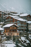 A cluster of wooden chalets with large windows is nestled on a snowy hillside. The buildings are covered with a layer of snow, and the background features a mountainous landscape with overcast skies.