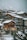 A cluster of wooden chalets with large windows is nestled on a snowy hillside. The buildings are covered with a layer of snow, and the background features a mountainous landscape with overcast skies.