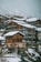 A cluster of wooden chalets with large windows is nestled on a snowy hillside. The buildings are covered with a layer of snow, and the background features a mountainous landscape with overcast skies.