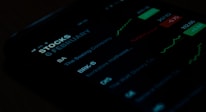 A close-up view of a smartphone screen displaying stock market information. The screen shows stock symbols such as BA and BRK-B, along with company names like The Boeing Company and Berkshire Hathaway. There are also green and red charts alongside figures indicating stock performance.
