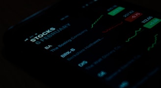 A close-up view of a smartphone screen displaying stock market information. The screen shows stock symbols such as BA and BRK-B, along with company names like The Boeing Company and Berkshire Hathaway. There are also green and red charts alongside figures indicating stock performance.