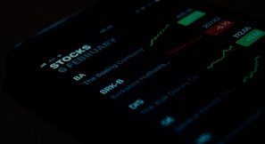 A close-up view of a smartphone screen displaying stock market information. The screen shows stock symbols such as BA and BRK-B, along with company names like The Boeing Company and Berkshire Hathaway. There are also green and red charts alongside figures indicating stock performance.