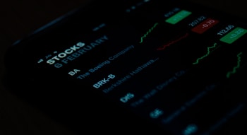 A close-up view of a smartphone screen displaying stock market information. The screen shows stock symbols such as BA and BRK-B, along with company names like The Boeing Company and Berkshire Hathaway. There are also green and red charts alongside figures indicating stock performance.