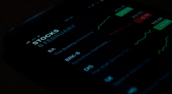 A close-up view of a smartphone screen displaying stock market information. The screen shows stock symbols such as BA and BRK-B, along with company names like The Boeing Company and Berkshire Hathaway. There are also green and red charts alongside figures indicating stock performance.