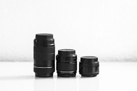 Three camera lenses of varying sizes are arranged side by side on a smooth, reflective surface against a plain white background. The tallest lens is on the left, with two progressively shorter lenses to the right. The lenses are black and cylindrical, featuring ridged focus rings and caps.