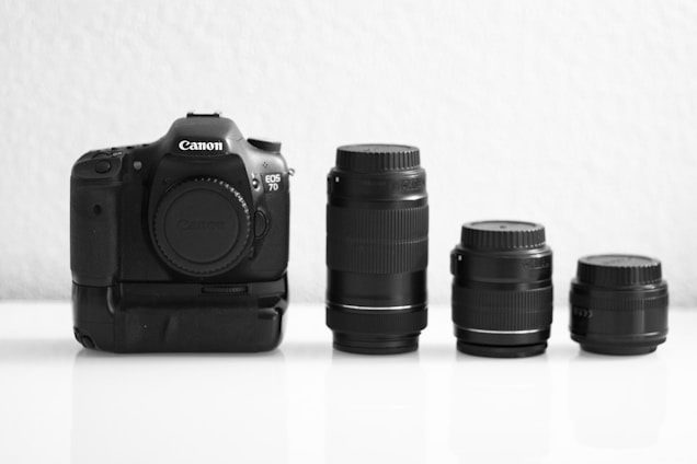 A Canon EOS 7D camera body with a battery grip is placed next to three interchangeable lenses of varying sizes on a white surface. The background is a plain, light-colored wall.
