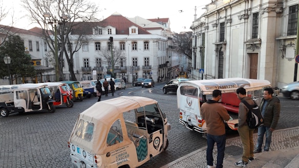 A cobblestone street lined with white buildings featuring red roofs, where multiple tuk-tuks are parked. A few people are gathered near one of the tuk-tuks, while others walk along the street. Bare trees and a clear sky are also visible.