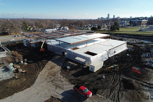 Aerial view of a construction site featuring a partially built structure with a large roof and steel framework. The ground is covered in dirt with visible tire tracks and construction equipment, including a crane and a red vehicle. Surrounding the site are urban elements, such as nearby roads, residential houses, and a few larger buildings in the distance.