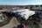 Aerial view of a construction site featuring a partially built structure with a large roof and steel framework. The ground is covered in dirt with visible tire tracks and construction equipment, including a crane and a red vehicle. Surrounding the site are urban elements, such as nearby roads, residential houses, and a few larger buildings in the distance.