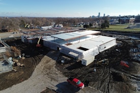 Aerial view of a construction site featuring a partially built structure with a large roof and steel framework. The ground is covered in dirt with visible tire tracks and construction equipment, including a crane and a red vehicle. Surrounding the site are urban elements, such as nearby roads, residential houses, and a few larger buildings in the distance.