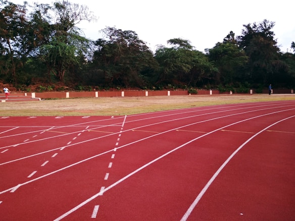 A running track with several lanes marked by white lines curves gently in the foreground. The track is red and appears well-maintained. In the background, there is a grassy area bordered by tall trees and a low brick wall. Two people are visible, one walking and another sitting, providing a sense of scale.