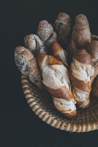 A wicker basket contains a variety of freshly baked crusty baguettes, some sprinkled with sesame and poppy seeds, arranged neatly against a dark background.