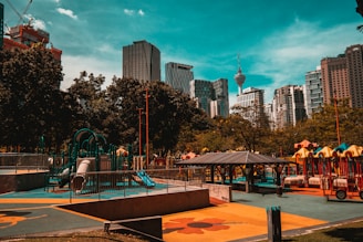 An urban playground with colorful equipment, surrounded by trees and high-rise buildings in the background. The structures include slides, climbing frames, and a pavilion. The sky is clear with a turquoise hue.