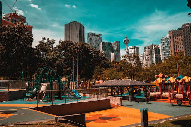 An urban playground with colorful equipment, surrounded by trees and high-rise buildings in the background. The structures include slides, climbing frames, and a pavilion. The sky is clear with a turquoise hue.