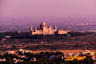 A grand palace-like structure with multiple domes and turrets stands amidst a sprawling, arid landscape. The architecture is intricate and majestic, set against a backdrop of a vast, slightly hazy cityscape. Sparse vegetation surrounds the building, with smaller structures scattered in the foreground, indicating a populated area.