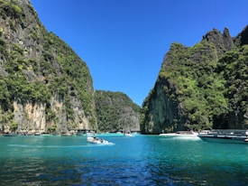 A picturesque scene featuring clear blue waters flanked by tall, verdant cliffs under a bright blue sky. Several boats are seen moving through the water, suggesting a lively and adventurous atmosphere.
