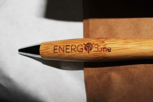A bamboo pen engraved with the text 'ENERG3.me' is placed on a brown surface, with a metal tip visible. The grain and texture of the bamboo give it a natural look.