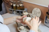 Hands are holding a small container, possibly of cosmetic or skincare product, surrounded by various glass bottles with liquid, likely oils, and other containers on a table. There's a decorative sign with the words 'be Joy' in the background.