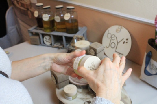 Hands are holding a small container, possibly of cosmetic or skincare product, surrounded by various glass bottles with liquid, likely oils, and other containers on a table. There's a decorative sign with the words 'be Joy' in the background.