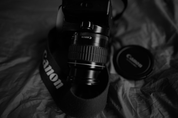 A digital camera with a Canon lens is positioned on a fabric surface. A branded camera strap is wrapped around the body, and a lens cap is placed nearby. The image is in black and white, highlighting the textures and shapes of the objects.