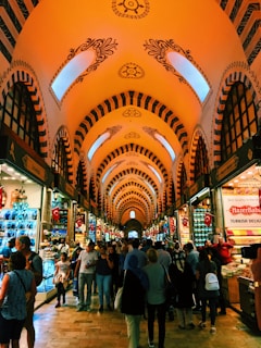 An indoor market with a high, elaborately decorated ceiling. Stalls on either side display various goods including Turkish delights and souvenirs. A crowd of people moves through the aisle, engaging in shopping and conversation. Bright lighting and colorful items create a lively atmosphere.