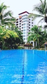 A luxurious outdoor swimming pool set against a backdrop of a multi-story building surrounded by tall palm trees. The water in the pool is vibrant blue, reflecting the sky and creating a serene atmosphere. Lush greenery surrounds the area, enhancing the tropical resort vibe.