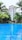 A luxurious outdoor swimming pool set against a backdrop of a multi-story building surrounded by tall palm trees. The water in the pool is vibrant blue, reflecting the sky and creating a serene atmosphere. Lush greenery surrounds the area, enhancing the tropical resort vibe.