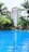 A luxurious outdoor swimming pool set against a backdrop of a multi-story building surrounded by tall palm trees. The water in the pool is vibrant blue, reflecting the sky and creating a serene atmosphere. Lush greenery surrounds the area, enhancing the tropical resort vibe.