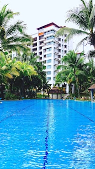 A luxurious outdoor swimming pool set against a backdrop of a multi-story building surrounded by tall palm trees. The water in the pool is vibrant blue, reflecting the sky and creating a serene atmosphere. Lush greenery surrounds the area, enhancing the tropical resort vibe.