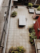 An aerial view of a tiled courtyard featuring a central table and chair, surrounded by potted ferns near the edges. A bicycle leans against a railing on the left, and there is a coil of hose in the upper corner. The courtyard is bordered by walls, one of which is partially covered in red and brick, with additional seating areas nearby.