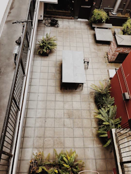 An aerial view of a tiled courtyard featuring a central table and chair, surrounded by potted ferns near the edges. A bicycle leans against a railing on the left, and there is a coil of hose in the upper corner. The courtyard is bordered by walls, one of which is partially covered in red and brick, with additional seating areas nearby.