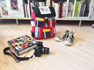 A colorful backpack with multiple pockets sits on a light wooden floor. Next to it, a pair of gray high-top sneakers is placed with shoelaces untied. A photography book, titled 'The Photo Book', lies beside a black DSLR camera with a strap. The background features a bookshelf filled with books of various sizes and colors.