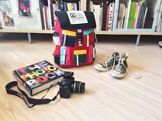 A colorful backpack with multiple pockets sits on a light wooden floor. Next to it, a pair of gray high-top sneakers is placed with shoelaces untied. A photography book, titled 'The Photo Book', lies beside a black DSLR camera with a strap. The background features a bookshelf filled with books of various sizes and colors.