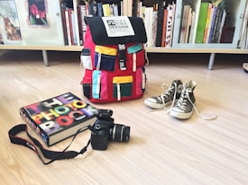A colorful backpack with multiple pockets sits on a light wooden floor. Next to it, a pair of gray high-top sneakers is placed with shoelaces untied. A photography book, titled 'The Photo Book', lies beside a black DSLR camera with a strap. The background features a bookshelf filled with books of various sizes and colors.