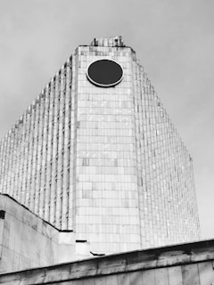 A large, modernist building with a distinctive pyramid-like shape, featuring a circular black emblem near the top. The structure is covered with numerous rectangular tiles, creating a textured facade. The architecture gives a sense of monumentalism and simplicity.